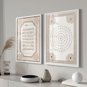 May include: Two framed Islamic art prints with white frames. The prints feature Arabic calligraphy in gold and beige tones. One print displays text, while the other has a circular design. The artwork is displayed on a light gray wall.