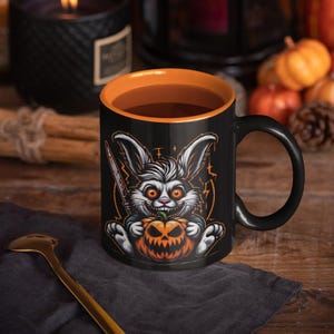 Könnte beinhalten: Schwarze Kaffeetasse mit orangefarbenem Rand, auf der ein Cartoon-Kaninchen abgebildet ist, das eine Halloween-Kürbis hält. Das Kaninchen hat einen bedrohlichen Ausdruck und hält ein blutiges Messer.