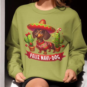 May include: Green sweatshirt featuring a cartoon dachshund wearing a sombrero and a festive sweater. The design includes cacti and the text "FELIZ NAVI-DOG" on a red banner. A fun, holiday-themed graphic.