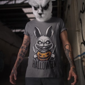 Könnte beinhalten: Ein graues T-Shirt mit einem Motiv eines weißen Kaninchens, das einen Kürbis mit einem grinsenden Gesicht und zwei Messer hält. Unter dem Kaninchen steht der Text "HALLOWEEN".