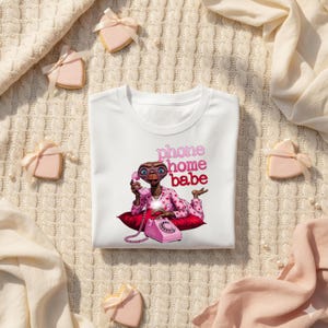 Op de afbeelding: Wit T-shirt met een afbeelding van E.T. die een roze telefoon vasthoudt, met de woorden "phone home babe" in roze. E.T. draagt een roze outfit met hartjespatroon en ligt op een rood kussen. Het shirt is omringd door koekjes en stof.