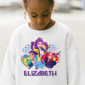 Może przedstawiać: Biała bluza z kolorową grafiką przedstawiającą trzy animowane postacie żeńskie, niebieskie stworzenie i imię "ELIZABETH" w kolorze fioletowym. Projekt jest umieszczony na środku przodu bluzy.