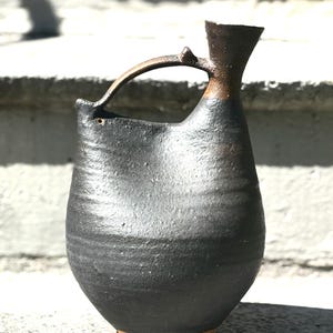 May include: A dark charcoal-colored ceramic pitcher with a textured, matte finish. The vessel features a rounded body, a narrow neck, and a single arched handle that connects the top of the neck to the side of the rim. The base is a light tan color.