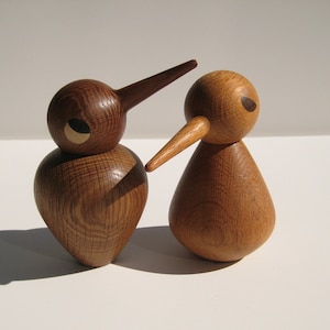 May include: Two wooden bird figurines with long beaks and rounded bodies. The birds are made of light brown wood with visible grain. One bird has a circular eye detail, and the other has a small, dark eye detail. The figurines are set against a white background.