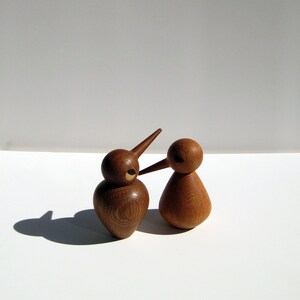 May include: Two wooden bird ornaments with long beaks and rounded bodies. The birds are crafted from light brown wood and feature a simple, minimalist design. One bird is taller than the other, and they are positioned close together.