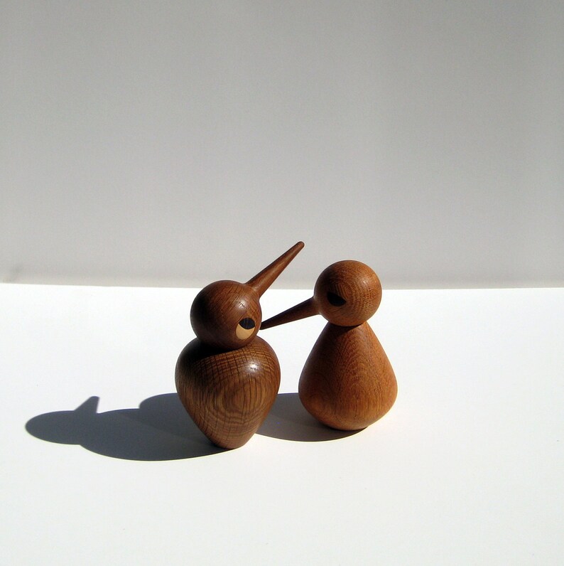 May include: Two wooden bird ornaments with long beaks and rounded bodies. The birds are crafted from light brown wood and feature a simple, minimalist design. One bird is taller than the other, and they are positioned close together.