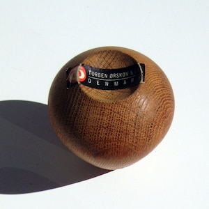 May include: A small, rounded wooden bowl with a textured surface and a dark opening. The bowl is light brown with a label that reads "TORBEN &Oslash;RSKOV & DENMARK". The bowl is likely a decorative item.