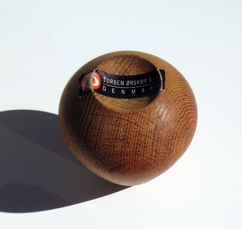 May include: A small, rounded wooden bowl with a textured surface and a dark opening. The bowl is light brown with a label that reads "TORBEN &Oslash;RSKOV & DENMARK". The bowl is likely a decorative item.