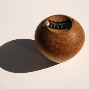 May include: A small, round wooden bowl with a smooth, polished finish. The bowl is made of light brown wood and has a circular indentation in the centre. A black and white label with text is affixed. The bowl casts a shadow on the white surface.