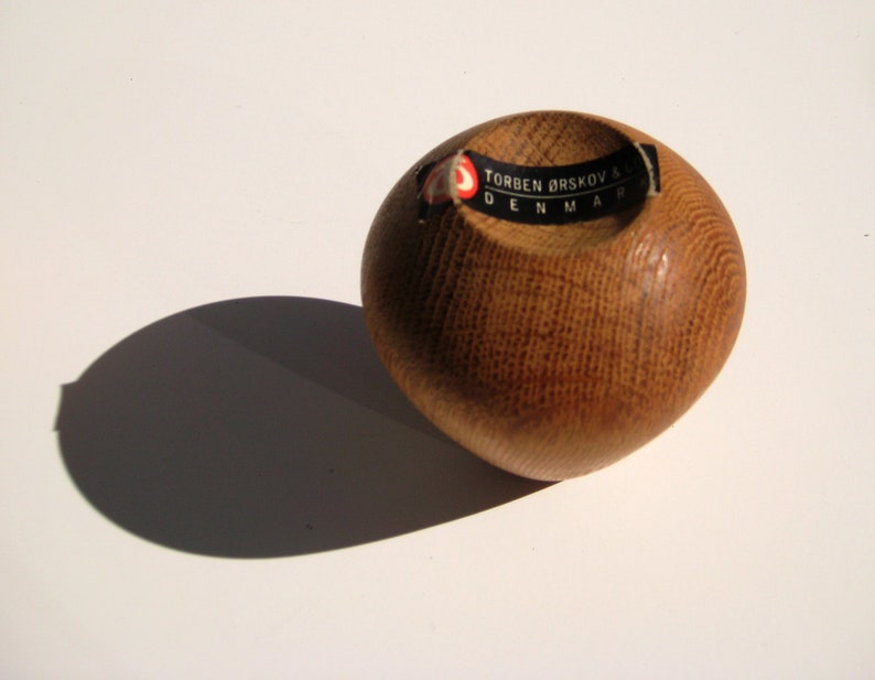 May include: A small, round wooden bowl with a smooth, polished finish. The bowl is made of light brown wood and has a circular indentation in the centre. A black and white label with text is affixed. The bowl casts a shadow on the white surface.