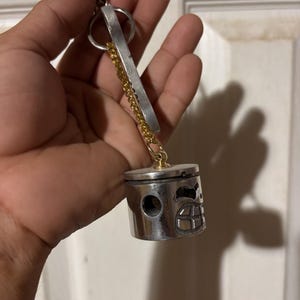 May include: A silver-colored piston keychain with a gold chain and a silver bar. The piston has a detailed face design. The keychain is attached to a silver clasp.
