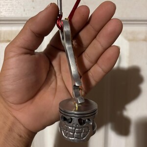 Op de afbeelding: Een zilverkleurige metalen sleutelhanger met een zuigervormige bedel met een grijnzend gezichtsontwerp. De sleutelhanger bevat een rode karabijnhaak voor eenvoudige bevestiging. De bedel heeft een gedetailleerde, industriële uitstraling.