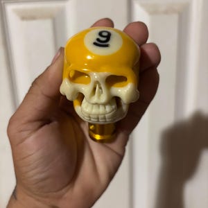 Puede incluir: Un pomo de palanca de cambios con forma de calavera amarilla y blanquecina. La parte superior del pomo es una bola de billar amarilla con el número 9 en negro. La calavera tiene un diseño detallado y realista.