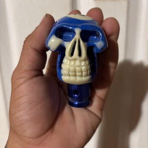 May include: A blue and white skull-shaped gear shift knob. The skull has a blue top with white accents and a cream-coloured jaw. The base of the knob is blue.