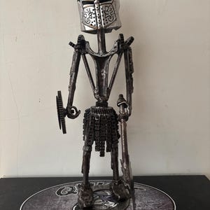 May include: A handcrafted metal sculpture of a knight, constructed from repurposed automotive parts. The knight features a piston helmet, gear shoulders, and a skirt made of metal links. It holds a spear and shield, standing on a base with text.