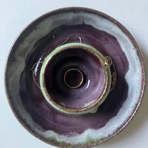 May include: A handmade ceramic jewelry dish with a deep purple and green glaze. A gold ring rests inside the small bowl, with a gold necklace draped over the edge. The dish is round with a wide rim.