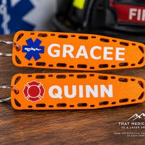 May include: Two orange keychains shaped like backboards with metal rings. The top keychain features a blue Star of Life emblem and the white text GRACEE. The bottom keychain features a red Maltese cross emblem and the white text QUINN.