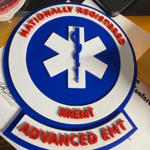 Könnte beinhalten: Ein rundes Emblem mit dem Text "NATIONALLY REGISTERED" in Rot, das einen blauen Kreis mit einem weißen Lebensstern-Symbol umgibt. Darunter steht der Text "NREMT ADVANCED EMT". Das Emblem ist weiß, blau und rot.