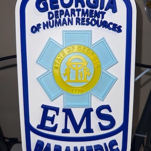 May include: A white, blue, and yellow rectangular sign with rounded corners. It features the text GEORGIA DEPARTMENT OF HUMAN RESOURCES, EMS, and PARAMEDIC in raised blue letters. A central light blue Star of Life emblem contains the State of Georgia seal.