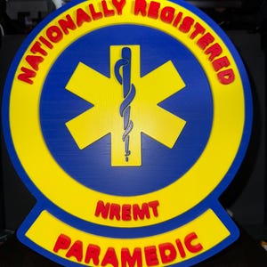 May include: A 3D-printed emblem featuring a blue circle with a yellow border and a yellow star of life. The words "NATIONALLY REGISTERED" and "PARAMEDIC" are in red, with "NREMT" in red below the star.