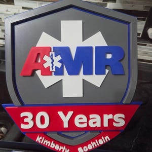 May include: A shield-shaped wall plaque featuring a grey background with a white Star of Life symbol. The letters AMR are in red and blue with a small Star of Life inside the A. Below, a red banner displays 30 Years and the name Kimberly Boehlein.