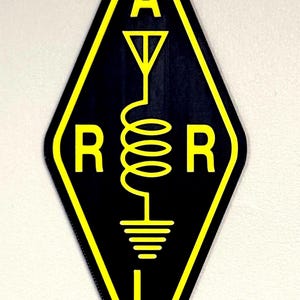 Könnte beinhalten: Schwarzes und gelbes rautenförmiges Schild mit den Buchstaben A, R und L sowie einem stilisierten Antennen-, Spulen- und Erdungssymbol. Das Schild ist wahrscheinlich für Funkamateure.