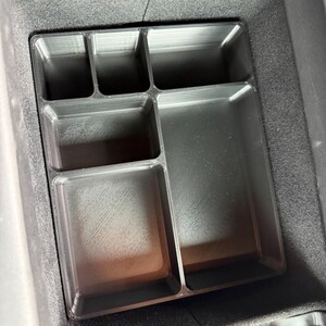 A black plastic center console organizer with five rectangular compartments, three small and two larger, sits inside a car's fuzzy-lined console storage.