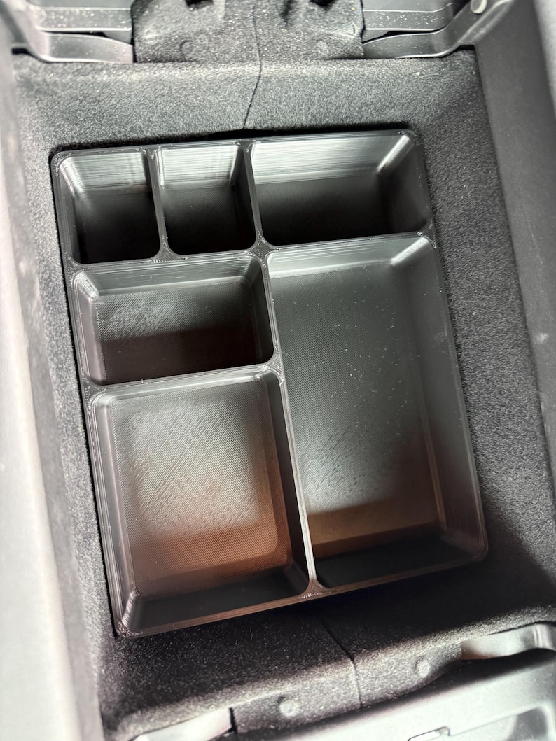 A black plastic center console organizer with five rectangular compartments, three small and two larger, sits inside a car's fuzzy-lined console storage.