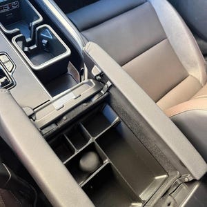 A black plastic center console organizer with multiple compartments is shown installed in a car's armrest storage, next to a brown and black car seat.