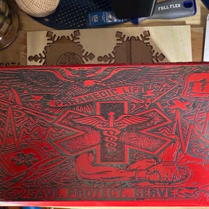Peut inclure: Une boîte rectangulaire rouge avec un design détaillé, avec les mots "PARAMEDIC LIFE" et "SAVE. PROTECT. SERVE." Le design comprend un symbole médical, un caducée et d'autres graphiques médicaux. La boîte est probablement pour les fournitures médicales.