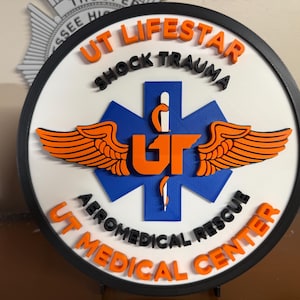 Puede incluir: Un letrero redondo con un borde negro que presenta el texto "UT LIFESTAR SHOCK TRAUMA AEROMEDICAL RESCUE UT MEDICAL CENTER". El letrero tiene un fondo blanco con un símbolo azul de la estrella de la vida y alas naranjas.