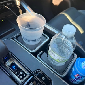 May include: A clear plastic cup with the text "Samford University Student Activities and Programs" sits in a car's cup holder, next to a water bottle and a container of gum. The cup holder is black and gray.