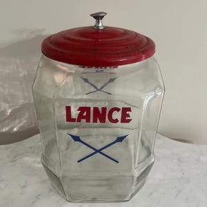 May include: A clear glass jar with a hexagonal shape and a red metal lid featuring a silver-colored knob. The front of the jar displays the red text LANCE above a blue graphic of two crossed spears. The glass has a wide opening and a sturdy base.