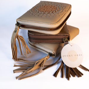May include: Three stacked, square leather wallets in beige and brown. The top wallet has a decorative stitched design. Each wallet has a zip closure and a tassel. A tag with the text "IVORY ORBS" hangs from one wallet.