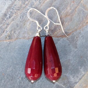 May include: A pair of dangle earrings with faceted, teardrop-shaped burgundy gemstones. The earrings have silver-coloured hooks and small silver beads at the top and bottom of each gemstone. The earrings are displayed against a grey stone surface.