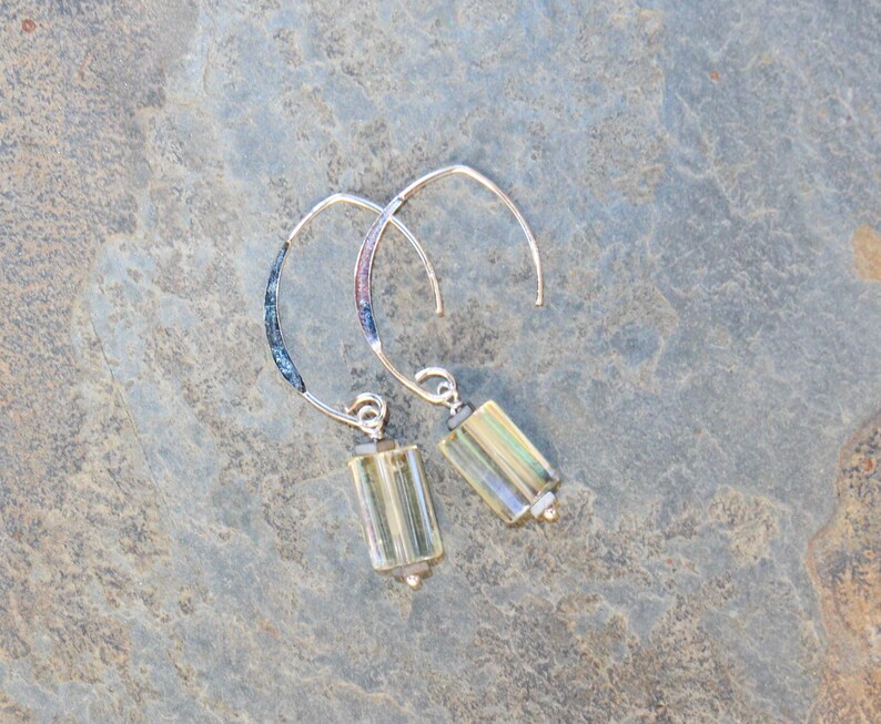 May include: A pair of silver hoop earrings with rectangular, light yellow-green beads. The hoops have a curved design with a textured, dark blue and purple section near the top. The earrings are displayed on a gray, textured surface.