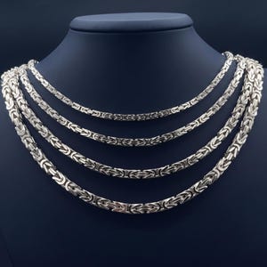 May include: Four silver-toned Byzantine link chains of varying thicknesses displayed on a dark neck form. Each chain features a complex, interlocking square-link pattern with a polished metallic finish, arranged in a cascading layered formation.