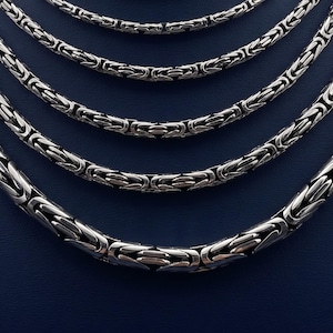 May include: A collection of five silver-toned Byzantine link chains arranged in a cascading semi-circular pattern. Each chain features a complex, woven metal design with interlocking segments that create a textured, cylindrical appearance.