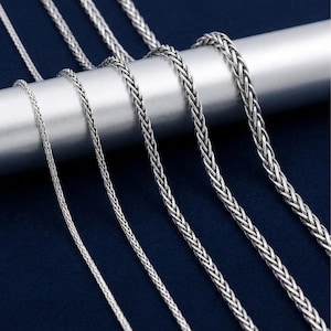 May include: A collection of five silver-toned wheat chains of varying thicknesses draped over a smooth, cylindrical bar. Each chain features a complex, braided link pattern with a metallic finish, showcasing a consistent design across all sizes.