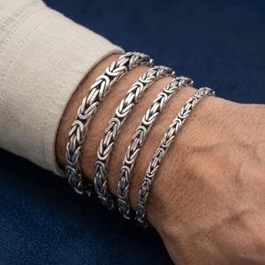 May include: Four silver bracelets featuring a Byzantine chain link pattern. The bracelets are arranged in a row, showcasing varying thicknesses of the interlocking metal links, which have a polished finish and dark oxidized detailing in the crevices.
