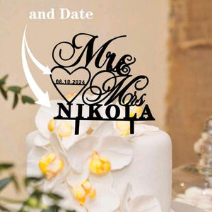 Peut inclure: Un cake topper noir avec l'inscription "Mrs & Mrs NIKOLA" et la date 08.10.2024, posé sur un gâteau blanc orné de fleurs blanches et jaunes. Le texte "Custom Name and Date" est au-dessus.