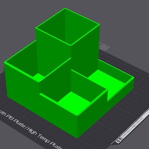 Könnte beinhalten: Ein leuchtend grüner, 3D-gedruckter Schreibtisch-Organizer mit mehreren Fächern. Das Design weist einen hohen, quadratischen Abschnitt und zwei niedrigere, rechteckige Abschnitte auf. Der Organizer steht auf einer dunkelgrauen Oberfläche.