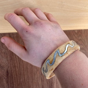 May include: A light-coloured wooden bracelet with a wavy silver metal inlay. The bracelet is worn on a wrist, showcasing the natural wood grain and the contrasting silver design. The bracelet is approximately 5 cm wide.