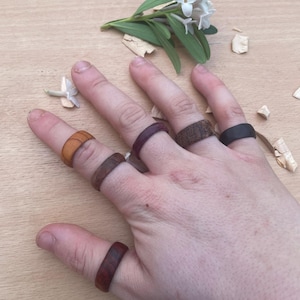 Puede incluir: Una colección de seis anillos de madera en varios tonos de marrón, desde el bronceado claro hasta el espresso oscuro y el morado intenso, usados en los dedos de una mano. Cada anillo presenta un diseño de banda lisa y redondeada de madera.
