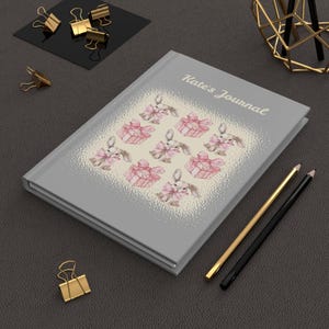 Könnte beinhalten: Ein graues Journal mit einem hellbeigen Hintergrund, auf dem sich ein sich wiederholendes Muster aus Hasen befindet, die rosa Geschenkboxen halten. Der Text "Kate's Journal" ist in Schwarz oben auf dem Cover gedruckt.