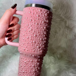Könnte beinhalten: Eine rosafarbene Edelstahl-Reisebecher mit Henkel und Deckel. Der Becher ist mit kleinen rosa Perlen bedeckt.