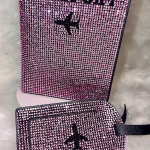 May include: A pink passport cover and matching luggage tag, both covered in rhinestones and featuring a black airplane design. The cover has the word "PASSPORT" written in black rhinestones.