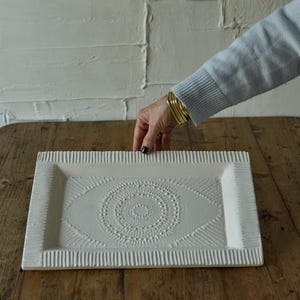 May include: A rectangular off-white tray featuring a central embossed circular mandala pattern and textured vertical line detailing along the raised edges. The tray has a matte finish and a shallow basin, showcasing a detailed relief design.