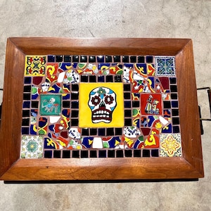 May include: A rectangular wooden serving tray with a colourful mosaic tile design. The mosaic features a central yellow square with a sugar skull, surrounded by various patterned tiles in red, blue, green, and black. The tray has metal handles with brown leather grips.