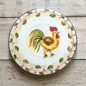 Puede incluir: Plato de cerámica decorativo con un diseño de gallo pintado a mano. El gallo es amarillo, marrón y verde, con una cresta roja. El plato tiene un centro color crema, un borde de hojas verdes y un borde azul.